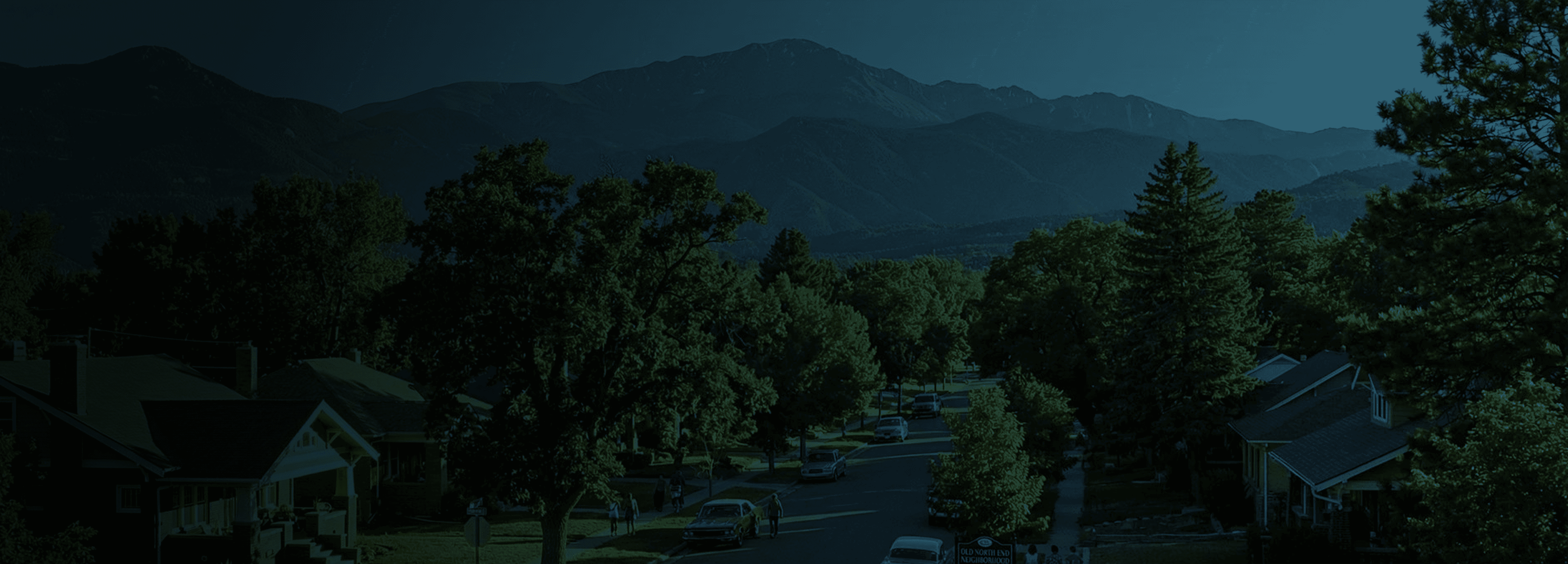 A tree-lined Colorado Springs neighborhood street with Pikes Peak rising behind it at dusk